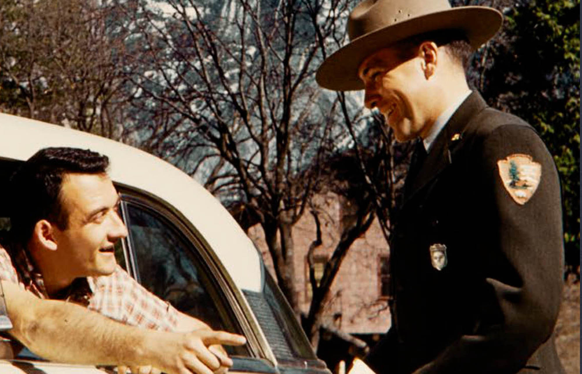 Man in an NPS uniform with broad brim hat and a silver shield shaped badge talks to a man leaning out of a car window.